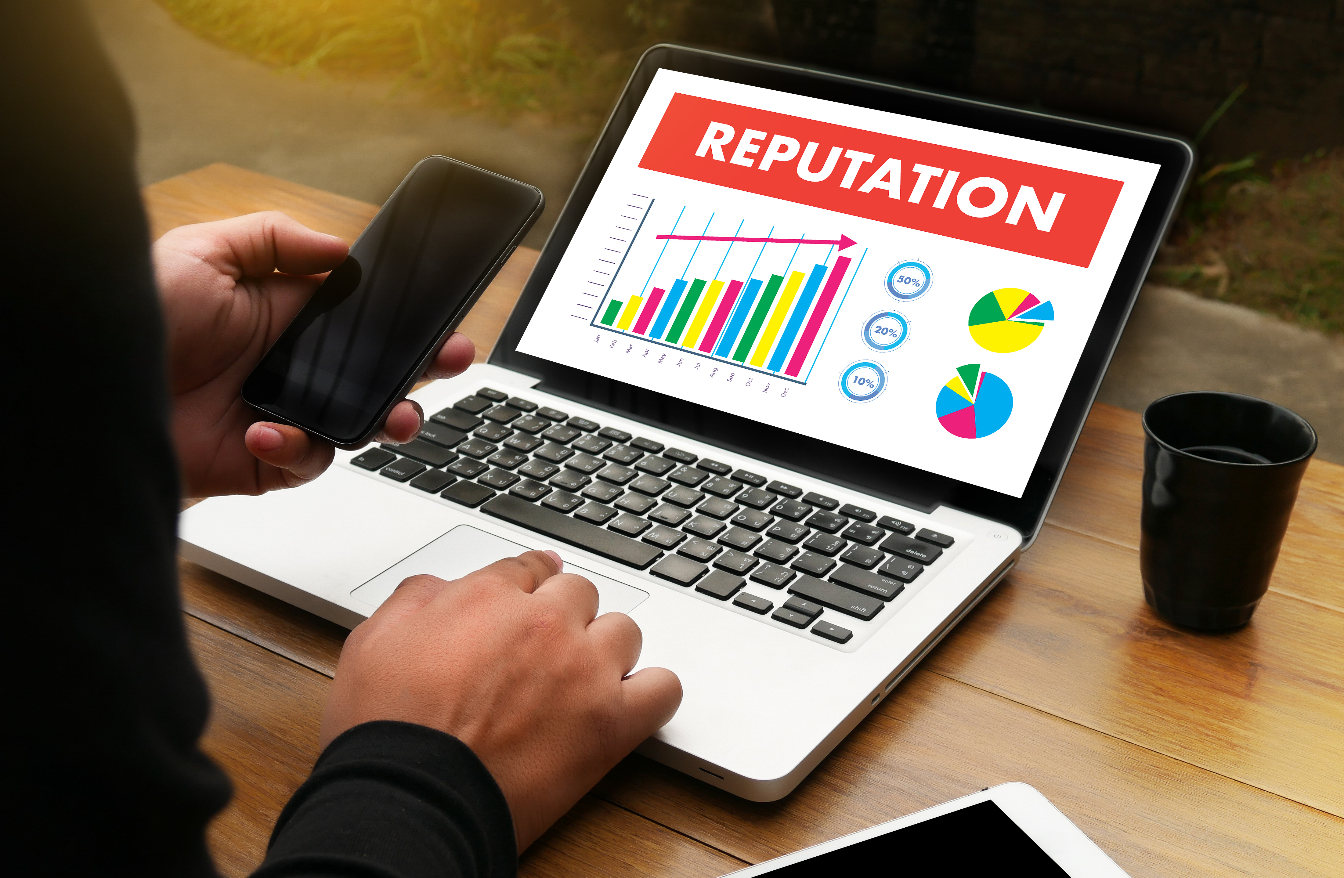 open laptop on desk with word 'reputation' on top of screen, cup on desk, person on left typing and holding phone